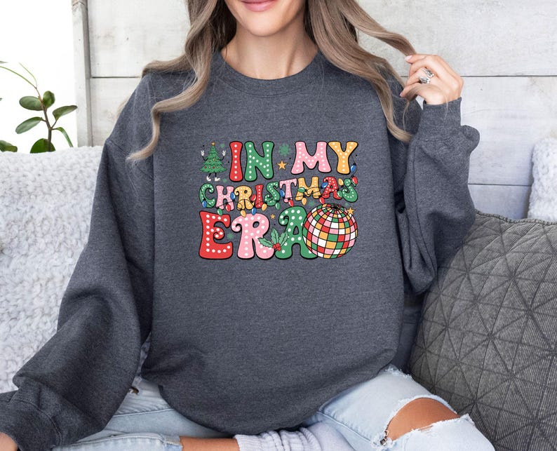 May include: Dark gray sweatshirt with the text "IN MY CHRISTMAS ERA" in colorful, retro-style lettering. The design includes a Christmas tree, a disco ball, and other festive elements, perfect for the holiday season.