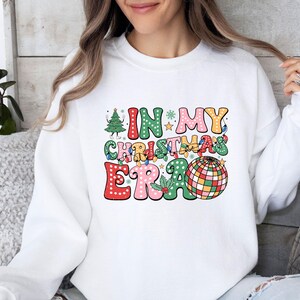 May include: White sweatshirt with the text "IN MY CHRISTMAS ERA" in colorful, retro-style lettering. The design includes a Christmas tree, string lights, and a disco ball, evoking a festive holiday theme.
