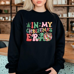 May include: Black crewneck sweatshirt with the text "IN MY CHRISTMAS ERA" in colorful, retro-style lettering. The design includes a Christmas tree, stars, lights, and a disco ball. A festive holiday-themed apparel item.