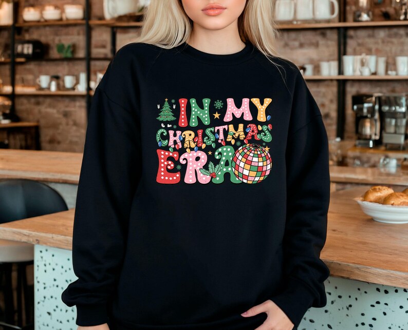 May include: Black crewneck sweatshirt with the text "IN MY CHRISTMAS ERA" in colorful, retro-style lettering. The design includes a Christmas tree, stars, lights, and a disco ball. A festive holiday-themed apparel item.