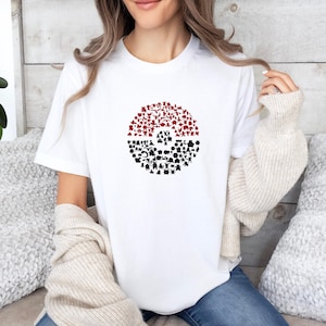 May include: A white short-sleeved t-shirt featuring a circular graphic on the chest. The graphic is shaped like a sphere, divided into a red top half and a black bottom half, composed of numerous small, distinct black and red character silhouettes.