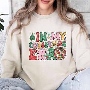 May include: A cream-colored sweatshirt with the phrase "IN MY CHRISTMAS ERA" in a colorful, retro font. The design includes a Christmas tree, lights, and a disco ball, evoking a festive holiday theme.