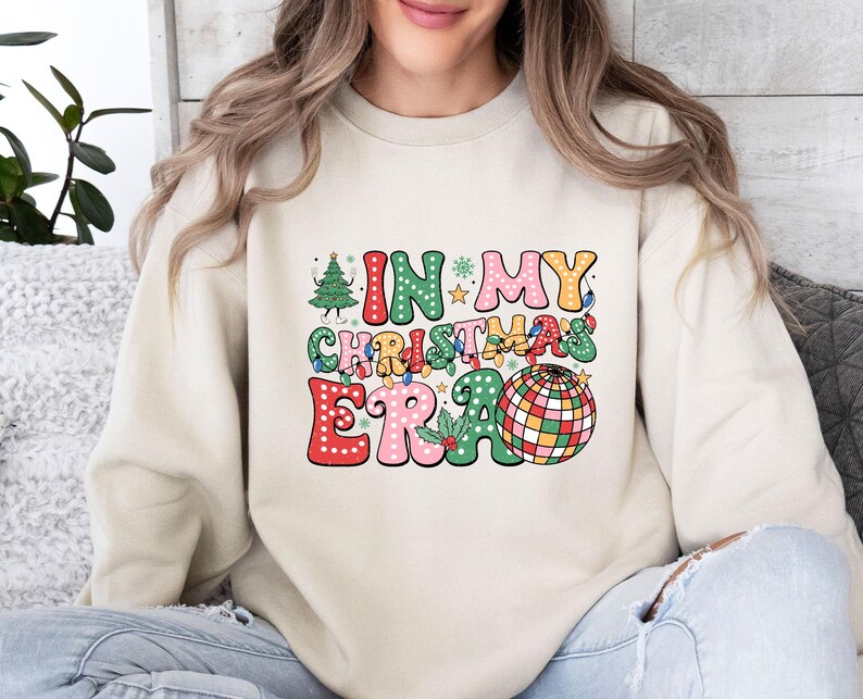 May include: A cream-colored sweatshirt with the phrase "IN MY CHRISTMAS ERA" in a colorful, retro font. The design includes a Christmas tree, lights, and a disco ball, evoking a festive holiday theme.
