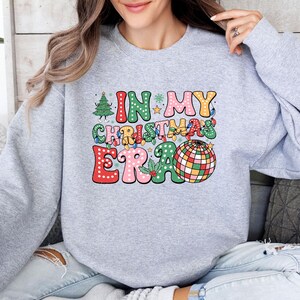 May include: A light gray sweatshirt with the text "IN MY CHRISTMAS ERA" in a colorful, retro design. The graphic includes a Christmas tree, string lights, and a disco ball. The sweatshirt is a comfortable, casual style.