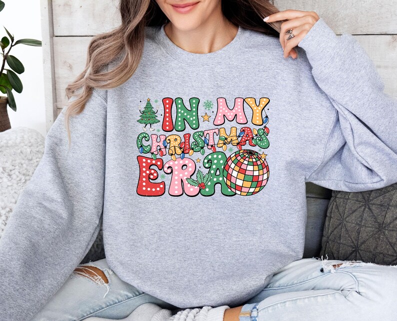 May include: A light gray sweatshirt with the text "IN MY CHRISTMAS ERA" in a colorful, retro design. The graphic includes a Christmas tree, string lights, and a disco ball. The sweatshirt is a comfortable, casual style.