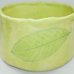 May include: A handmade, light green ceramic container with a leaf design. The cylindrical pot has a slightly irregular rim and a large, detailed green leaf painted on the side. The overall color is a soft, pale green.