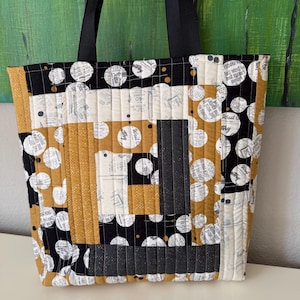 May include: A quilted tote bag with a patchwork design. The bag features black, white, and mustard yellow fabric with a polka dot and newspaper print. It has black handles and a rectangular shape.