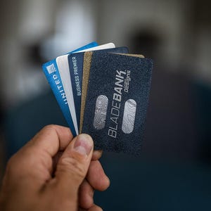 May include: A hand holding a stack of credit cards. The top card is black with the text "BLADEBANK" in white and the words "Design Credit" in smaller white text. The other cards are blue and white with the text "UNITED" and "BUSINESS PREMIER" visible.