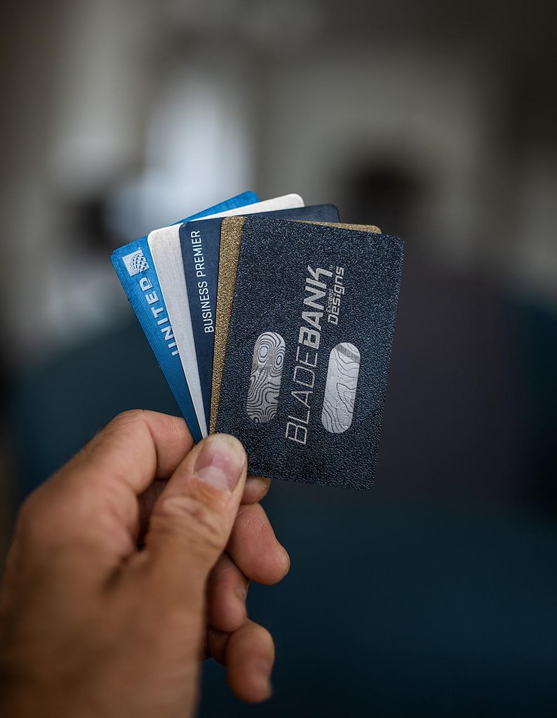 May include: A hand holding a stack of credit cards. The top card is black with the text "BLADEBANK" in white and the words "Design Credit" in smaller white text. The other cards are blue and white with the text "UNITED" and "BUSINESS PREMIER" visible.