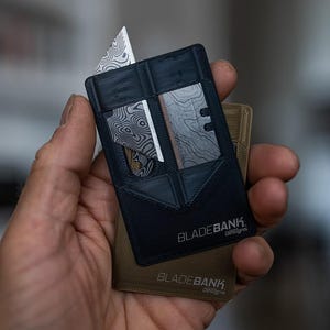 May include: A black card-sized multi-tool with a silver blade and the text "BLADEBANK. Designs" printed on it. The tool is being held in a hand.