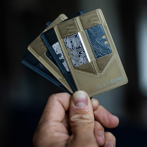 May include: A stack of four minimalist wallets, two are black and two are tan. The tan wallets have a textured surface and the text "The Bank" printed on the bottom right corner.
