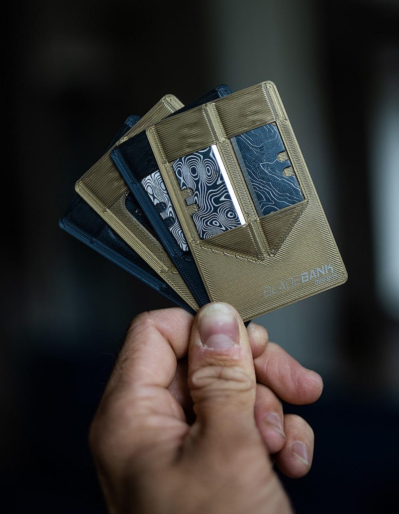 May include: A stack of four minimalist wallets, two are black and two are tan. The tan wallets have a textured surface and the text "The Bank" printed on the bottom right corner.