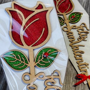 Puede incluir: Una rosa de madera con p&eacute;talos de purpurina roja y hojas de purpurina verde. La rosa est&aacute; unida a un tallo de madera con las palabras "Feliz Dia" escritas en &eacute;l.