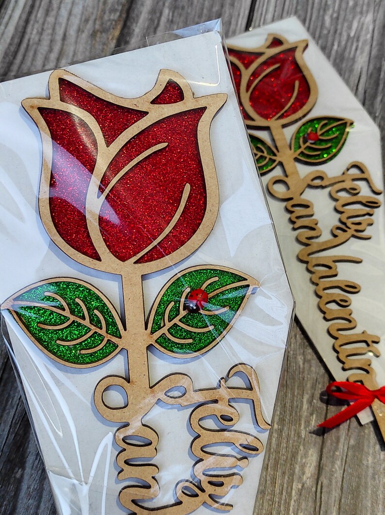 Puede incluir: Una rosa de madera con p&eacute;talos de purpurina roja y hojas de purpurina verde. La rosa est&aacute; unida a un tallo de madera con las palabras "Feliz Dia" escritas en &eacute;l.