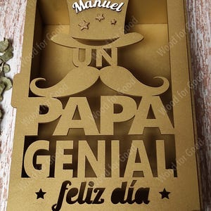 Może przedstawiać: Złote drewniane pudełko z wyciętym wzorem, na kt&oacute;rym widnieje napis "Un Papa Genial" i "Feliz dia". Pudełko ma mały otw&oacute;r na g&oacute;rze z wycięciem w kształcie cylindra i wąs&oacute;w. Na cylindrze widnieje napis "Manuel".