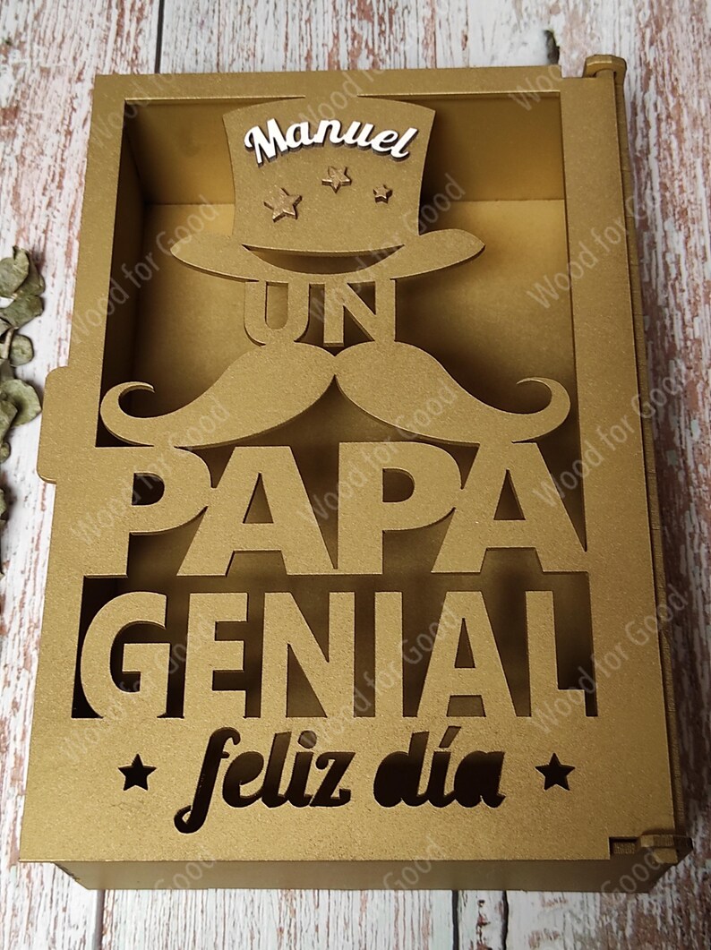 Może przedstawiać: Złote drewniane pudełko z wyciętym wzorem, na kt&oacute;rym widnieje napis "Un Papa Genial" i "Feliz dia". Pudełko ma mały otw&oacute;r na g&oacute;rze z wycięciem w kształcie cylindra i wąs&oacute;w. Na cylindrze widnieje napis "Manuel".
