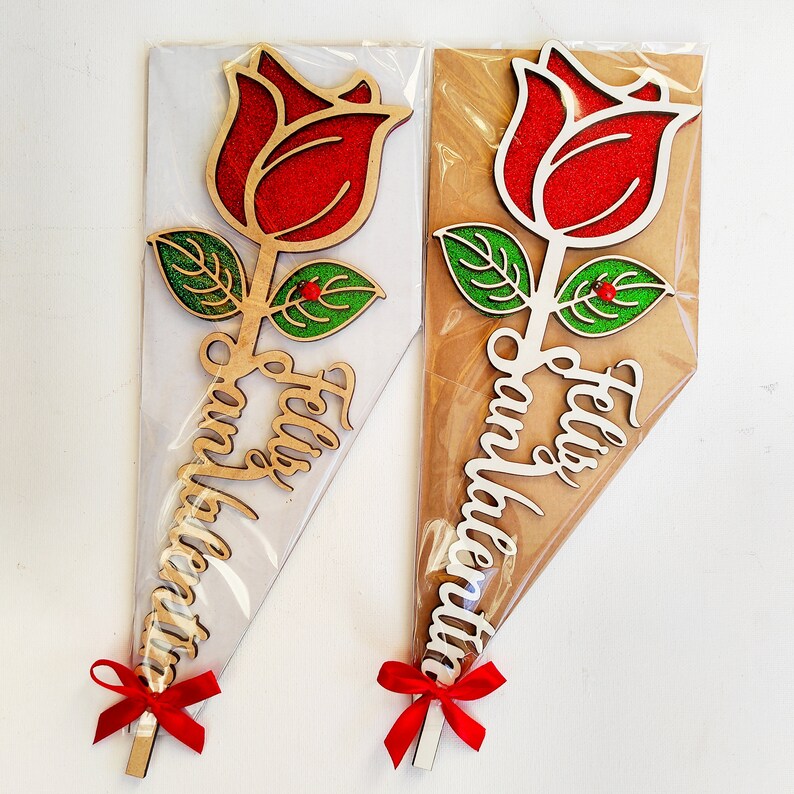Puede incluir: Dos ramos de rosas de madera con rosas rojas de purpurina y hojas verdes. Los ramos est&aacute;n envueltos en pl&aacute;stico transparente y tienen lazos de cinta roja. Los tallos est&aacute;n decorados con las palabras "Feliz San Valentin" en blanco.