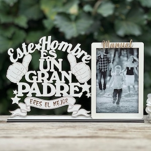 May include: White wooden sign with the text "Este Hombre Es Un Gran Padre Eres El Mejor" and two thumbs up. A white picture frame with the name "Manuel" at the top and a black and white photo of a family inside.