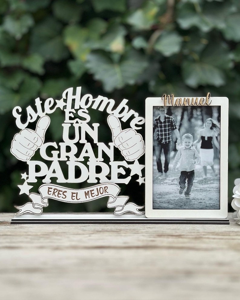May include: White wooden sign with the text "Este Hombre Es Un Gran Padre Eres El Mejor" and two thumbs up. A white picture frame with the name "Manuel" at the top and a black and white photo of a family inside.