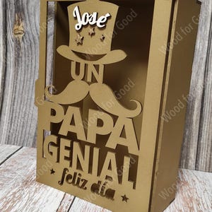 Może przedstawiać: Złote drewniane pudełko z wyciętym wzorem wąs&oacute;w, kapelusza i napisami "Un Papa Genial" i "Feliz Dia". Na kapeluszu widnieje imię "Jose".
