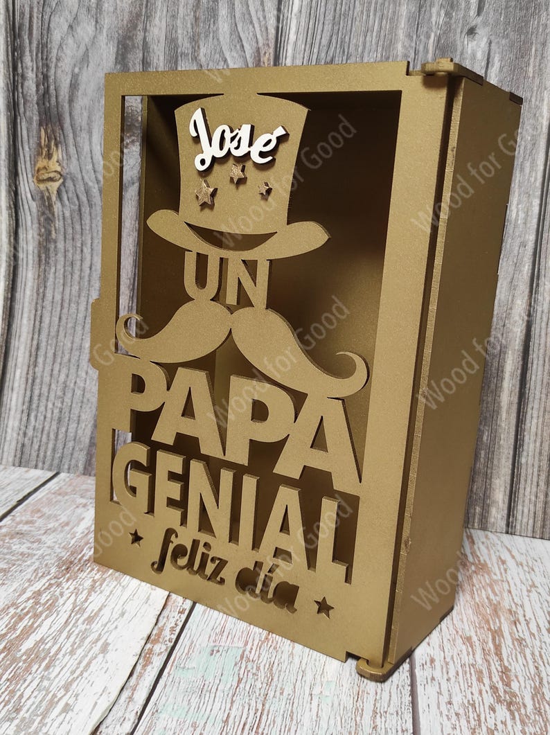 Może przedstawiać: Złote drewniane pudełko z wyciętym wzorem wąs&oacute;w, kapelusza i napisami "Un Papa Genial" i "Feliz Dia". Na kapeluszu widnieje imię "Jose".