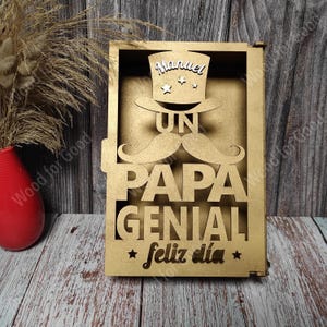 Może przedstawiać: Drewniany znak z wyciętym wzorem przedstawiającym cylinder, wąsy i słowa "Un Papa Genial Feliz Dia". Na cylindrze widnieje imię "Manuel".