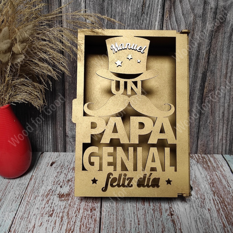 Może przedstawiać: Drewniany znak z wyciętym wzorem przedstawiającym cylinder, wąsy i słowa "Un Papa Genial Feliz Dia". Na cylindrze widnieje imię "Manuel".