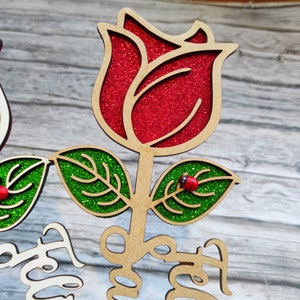 Puede incluir: Una rosa de madera con p&eacute;talos rojos de purpurina y hojas verdes de purpurina. Dos peque&ntilde;as mariquitas rojas est&aacute;n en las hojas. El tallo es marr&oacute;n y las palabras "You are" est&aacute;n escritas en el tallo.