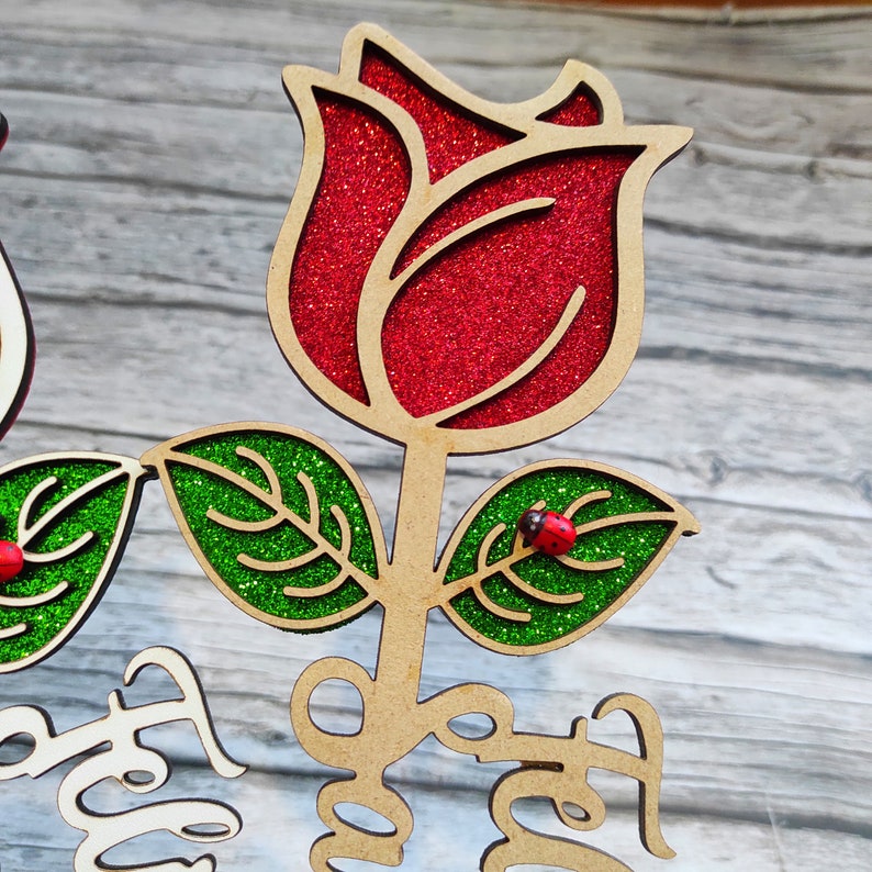 Puede incluir: Una rosa de madera con p&eacute;talos rojos de purpurina y hojas verdes de purpurina. Dos peque&ntilde;as mariquitas rojas est&aacute;n en las hojas. El tallo es marr&oacute;n y las palabras "You are" est&aacute;n escritas en el tallo.