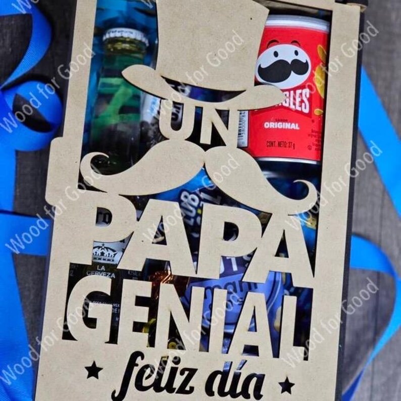 Może przedstawiać: Drewniana skrzynka z wyciętym wzorem, na kt&oacute;rym widnieje napis "Un Papa Genial" i "Feliz Dia" z wąsem i kapeluszem cylindrycznym. Skrzynka jest wypełniona r&oacute;żnymi przekąskami i napojami.