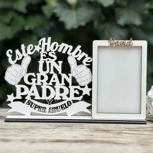 May include: White wooden picture frame with the text "Este Hombre Es Un Gran Padre Y Super Abuelo" and a thumbs up design. The frame has a smaller white frame inside with the name "David" written in brown.