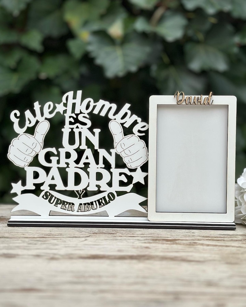 May include: White wooden picture frame with the text "Este Hombre Es Un Gran Padre Y Super Abuelo" and a thumbs up design. The frame has a smaller white frame inside with the name "David" written in brown.