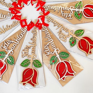 Puede incluir: Una colecci&oacute;n de adornos de madera con forma de rosa con el texto "Feliz San Valentin" escrito en ellos. Los adornos son rojos, verdes y blancos y est&aacute;n empaquetados en pl&aacute;stico transparente.