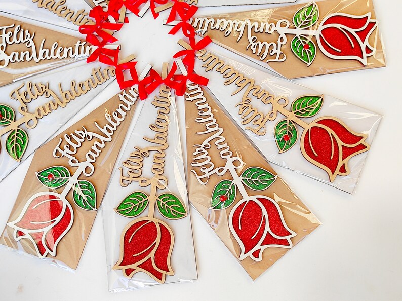 Puede incluir: Una colecci&oacute;n de adornos de madera con forma de rosa con el texto "Feliz San Valentin" escrito en ellos. Los adornos son rojos, verdes y blancos y est&aacute;n empaquetados en pl&aacute;stico transparente.