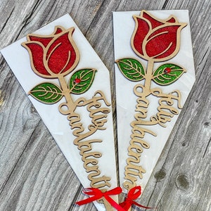 Puede incluir: Dos recortes de rosas de madera con rosas rojas de purpurina y hojas verdes de purpurina. Las rosas est&aacute;n unidas a palos y tienen cintas rojas atadas alrededor. Los palos tienen el texto "Feliz San Valentin" escrito en ellos.