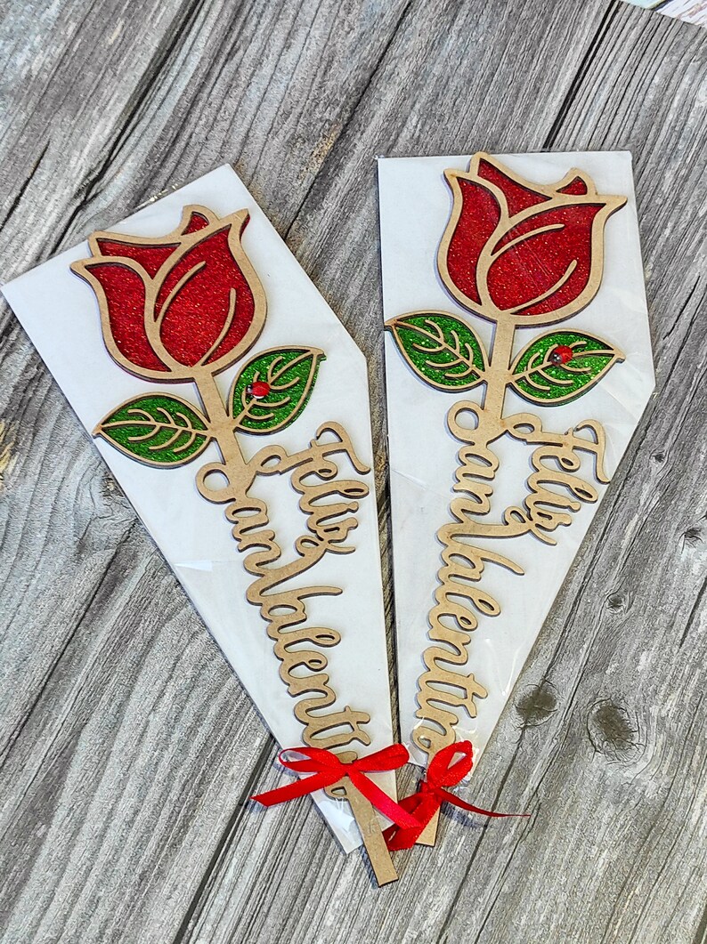 Puede incluir: Dos recortes de rosas de madera con rosas rojas de purpurina y hojas verdes de purpurina. Las rosas est&aacute;n unidas a palos y tienen cintas rojas atadas alrededor. Los palos tienen el texto "Feliz San Valentin" escrito en ellos.