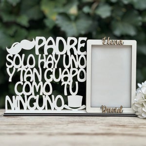 May include: White wooden picture frame with a cutout design that reads "Padre solo hay uno y tan guapo como tu ninguno". The frame has a small mustache cutout at the top. The frame is sitting on a wooden surface. The names "Elena" and "David" are written on the frame.