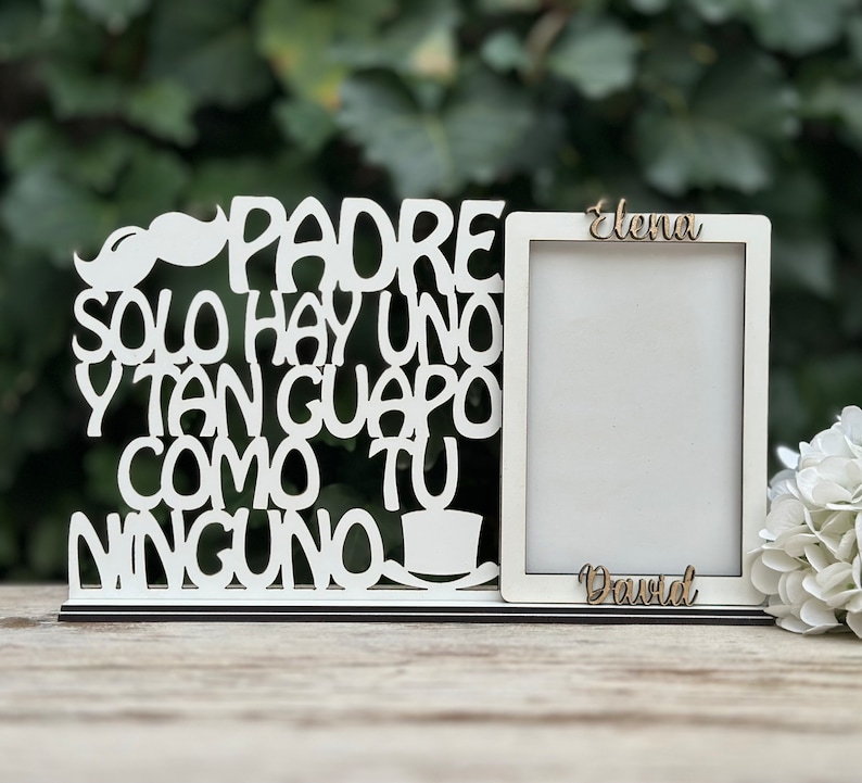May include: White wooden picture frame with a cutout design that reads "Padre solo hay uno y tan guapo como tu ninguno". The frame has a small mustache cutout at the top. The frame is sitting on a wooden surface. The names "Elena" and "David" are written on the frame.