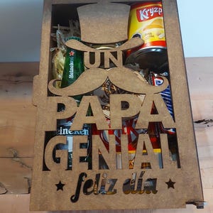 Może przedstawiać: Drewniana skrzynka z wyciętym wzorem, na kt&oacute;rym widnieje napis "Un Papa Genial Feliz Dia" z wąsem i kapeluszem. Skrzynka jest wypełniona r&oacute;żnymi przedmiotami, w tym piwem, chipsami i słodyczami.