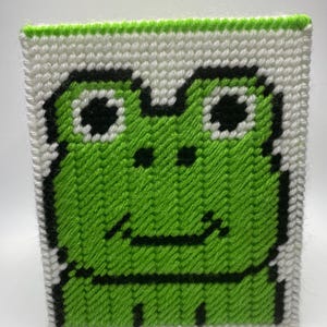 May include: A handmade, square tissue box cover featuring a pixelated frog design. The frog is primarily bright green with black outlines and eyes, set against a white background. The edges are trimmed with green yarn.