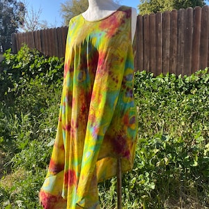 May include: A tie-dye tunic in shades of yellow, red, and blue is displayed on a mannequin. The sleeveless top has a round neckline and a flowing, asymmetrical hemline. The fabric appears soft and lightweight, suitable for warm weather.