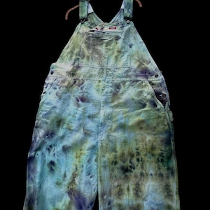 May include: Denim overalls featuring a tie-dye pattern in shades of light blue, sage green, and dark purple. The garment includes adjustable shoulder straps with metal buckles, a front bib pocket with a small red brand label, and side pockets.