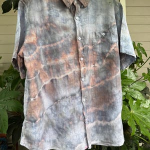 May include: A short-sleeved button-down shirt with a tie-dye pattern in shades of blue, gray, and peach. The shirt has a chest pocket and white buttons. The collar is button-down.