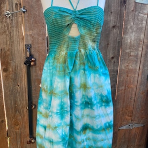 May include: A turquoise and blue tie-dye jumpsuit with a halter neckline and a keyhole cutout. The bodice is smocked, and the fabric has a flowing, lightweight appearance. The jumpsuit is displayed on a mannequin against a wooden fence.