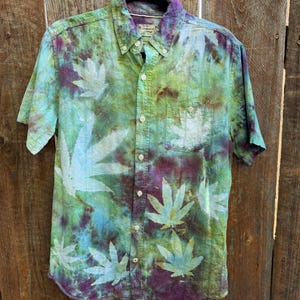 May include: A short-sleeved button-down shirt with a unique tie-dye pattern. The shirt features a blend of green, blue, and purple hues, with white leaf-shaped designs. It has a chest pocket and is hanging on a black hanger against a wooden background.