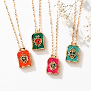 Peut inclure: Quatre colliers dorés avec des pendentifs rectangulaires orange, vert, rose et turquoise. Chaque pendentif présente un motif de cœur avec des détails d'œil et de flamme. Les colliers sont présentés sur une surface blanche avec de petites fleurs blanches.