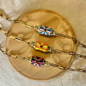 Peut inclure: Trois bracelets à chaîne dorés avec des pendentifs en forme d'enveloppe en émail coloré. Chaque pendentif présente un motif de cœur et est orné de strass. Les bracelets sont présentés sur un plateau en bois.
