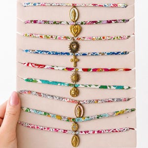 Peut inclure: Une collection de huit bracelets en ruban de tissu arborant des motifs floraux colorés dans des teintes variées. Chaque bracelet est orné d'une breloque en métal doré unique, incluant des icônes religieuses, un cœur, un soleil et une croix.