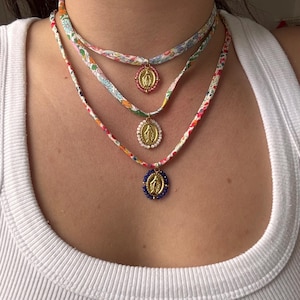Peut inclure: Trois colliers superposés avec des lanières en tissu à motifs floraux. Chaque collier comporte un pendentif doré avec une figure religieuse. Le pendentif supérieur a une bordure rose, le milieu une bordure de perles blanches et le bas une bordure de perles bleues.
