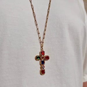 Peut inclure: Un collier doré composé d'une longue chaîne à maillons rectangulaires et d'un pendentif en forme de croix. La croix est ornée de pierres précieuses rectangulaires rouges, vertes et bleues, rehaussées de petites pierres claires sur métal doré.